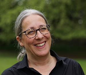 A woman with grey hair in a ponytail and tortoiseshell glasses wears a black shirt and sports a big friendly smile.