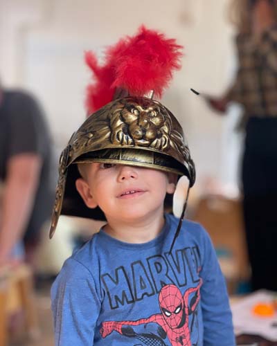 A child looks out from under a legionnaire's helmet on his head, which is falling over one eye