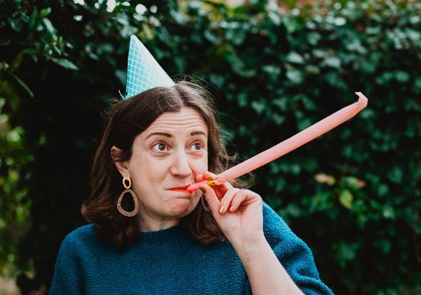 Leanne blows a party horn while wearing a party hat, standing in front of a bush.