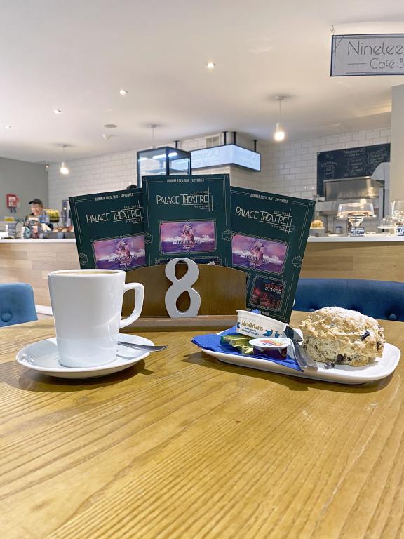 Coffee and scone on a café table with Palace Theatre brochures and Café Nineteen20 in the background.