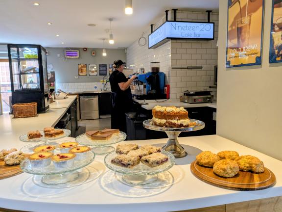 Café counter displaying cakes, scones and pastries, with a staff member preparing drinks behind the bar.