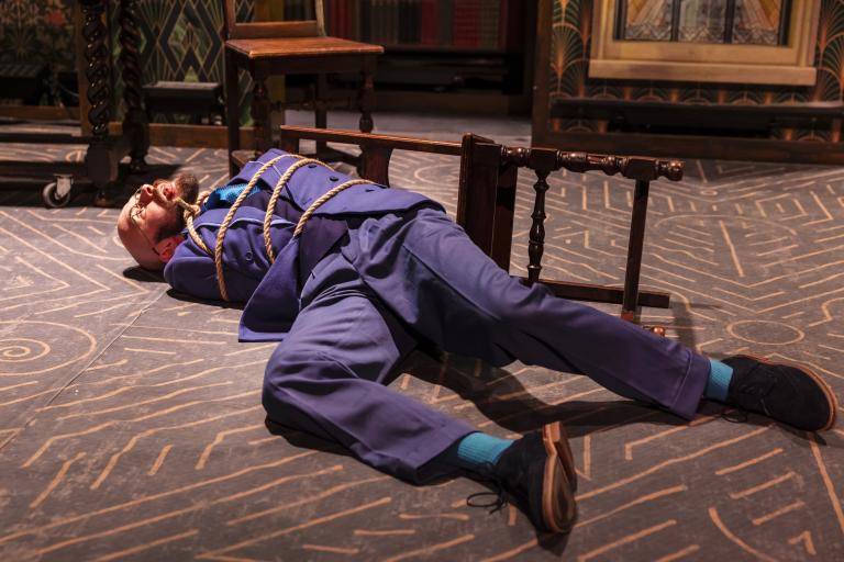 Production photo showing a performer in a blue suit tied to a fallen wooden chair with rope, lying on an art deco-style stage set. The scene features patterned flooring, dark wood furniture and bookshelves, suggesting a theatrical mystery or comedy production.