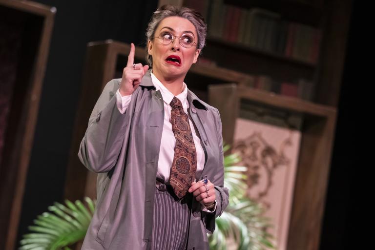 Production photo of a performer in a trench coat, striped trousers and glasses standing on stage with one finger raised. Bookshelves, framed artwork and plants in the background create the setting of a theatrical mystery or comedy production.