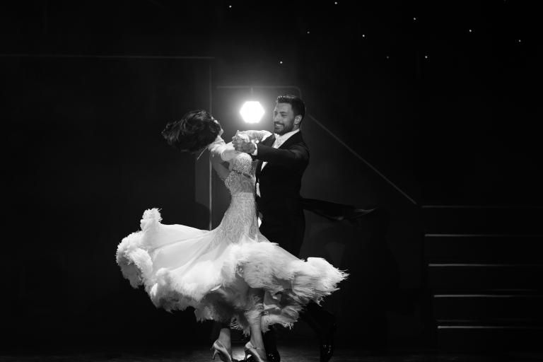 Black-and-white photograph of two ballroom dancers performing beneath a spotlight, with a flowing feather-trimmed gown creating a sense of movement on stage.