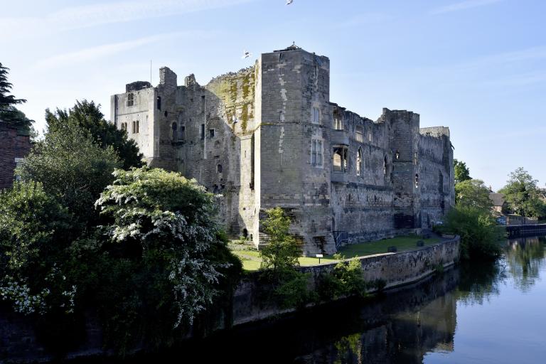 Newark Castle ruins beside the River Trent, surrounded by trees and reflected in the water beneath a clear blue sky.