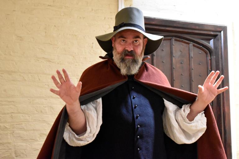 Costumed historical interpreter dressed in 17th-century clothing, including a wide-brimmed hat and cloak, standing inside a historic building. The interpreter gestures with raised hands while demonstrating life during the English Civil Wars, with brick walls and a wooden doorway visible in the background.
