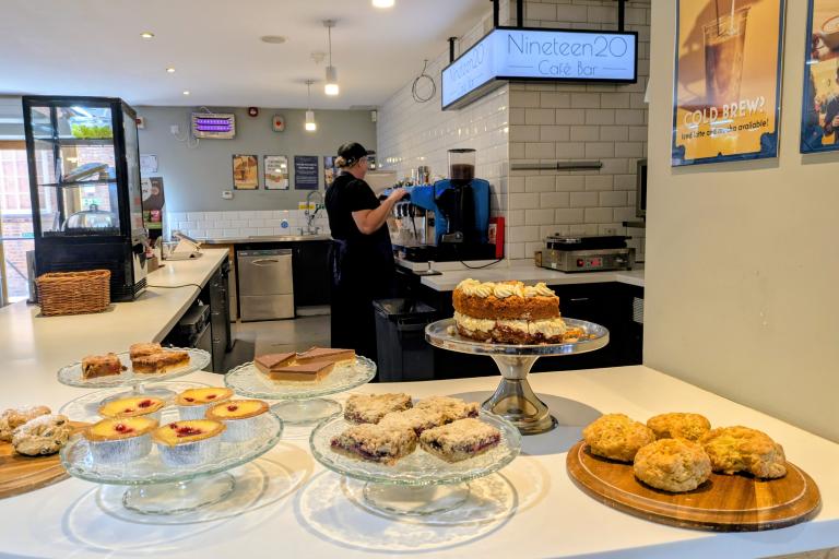Café counter displaying cakes, scones and pastries, with a staff member preparing drinks behind the bar.