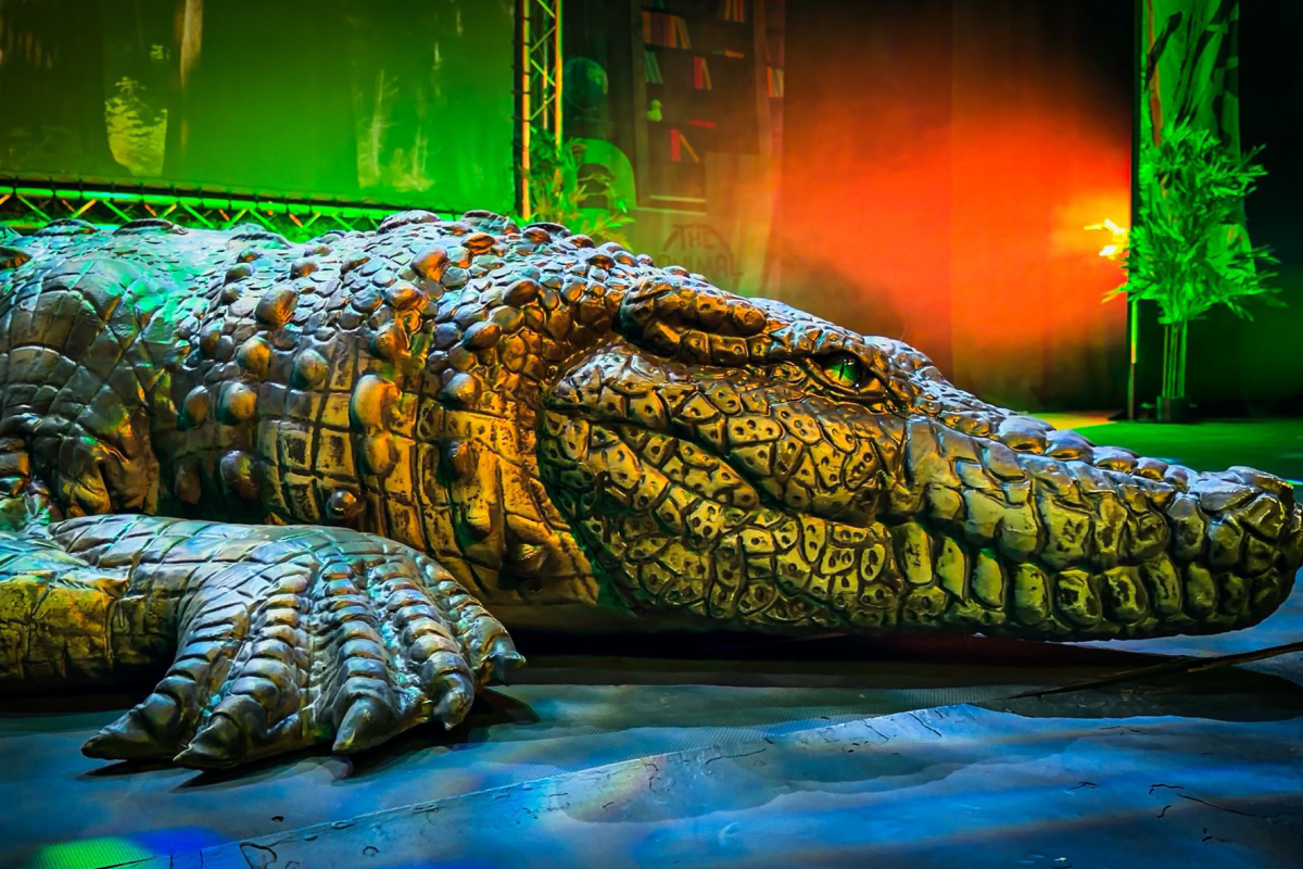 Large crocodile model on stage beneath colourful theatrical lighting and special effects during an animal-themed live show.