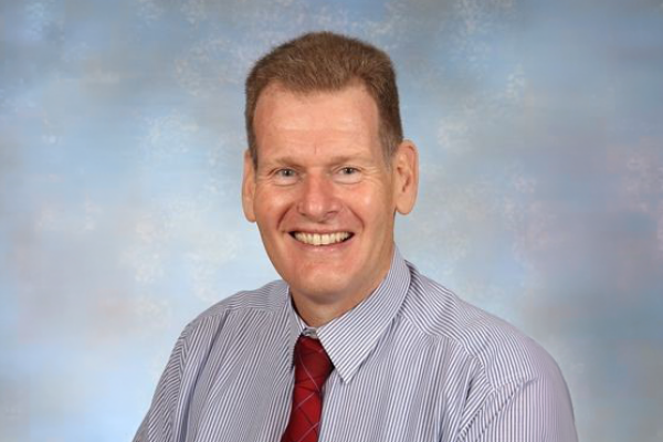 Professional headshot of a person wearing a blue striped shirt and red tie against a light blue studio background. The portrait is cropped from the chest up and shows the individual facing the camera.