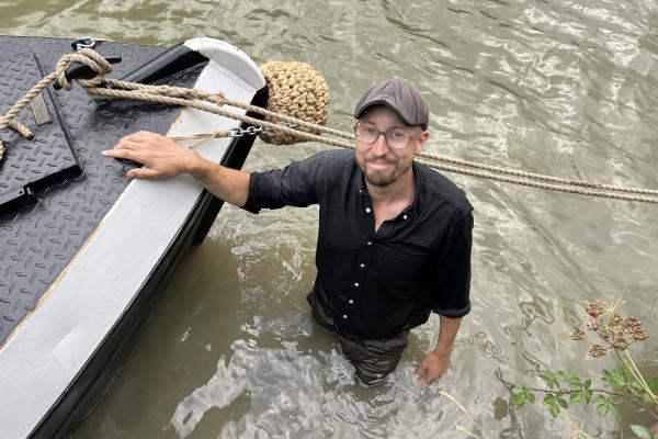 Person standing in a river next to a moored narrowboat, holding the bow and looking towards the camera.
