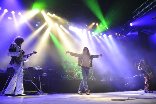 Live rock band performing on stage beneath colourful concert lighting, with a vocalist at centre stage and musicians playing around them.