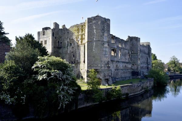 Newark Castle ruins beside the River Trent, surrounded by trees and reflected in the water beneath a clear blue sky.