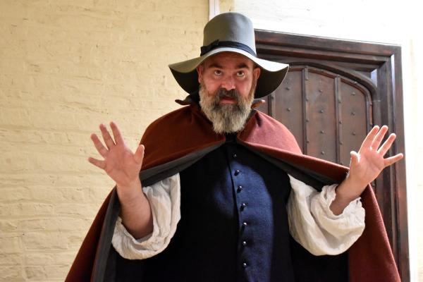 Costumed historical interpreter dressed in 17th-century clothing, including a wide-brimmed hat and cloak, standing inside a historic building. The interpreter gestures with raised hands while demonstrating life during the English Civil Wars, with brick walls and a wooden doorway visible in the background.
