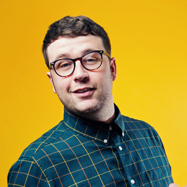 Head-and-shoulders promotional portrait of comedian Chris Washington wearing round glasses and a dark blue check-patterned shirt, set against a bright yellow background.