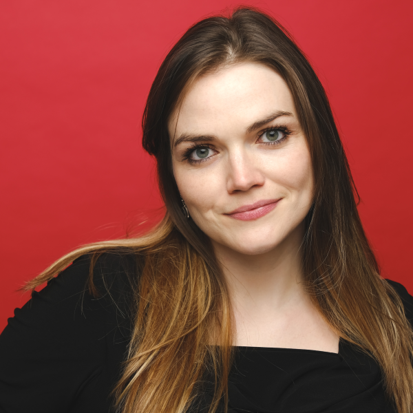 Head-and-shoulders promotional portrait of comedian Naomi Cooper, with long hair wearing a black top, photographed against a bright red background.