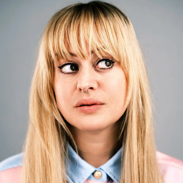 Head-and-shoulders promotional portrait of comedian Sammy Dobson with long blonde hair and a septum piercing, wearing a pastel collared shirt against a plain grey background.